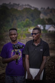 Two individuals stand outdoors, one holding a camera mounted on a stabilizer and the other holding a laptop. They are dressed casually, with one wearing a purple shirt and the other a black polo shirt. The background features blurred greenery and a house.