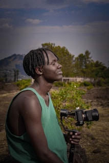 A person with closed eyes is holding a camera mounted on a gimbal. The background features lush greenery and distant hills under a partly cloudy sky.