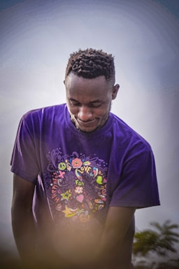 A person wearing a purple shirt with colorful graphic designs stands outdoors. The design includes words like 'love', 'fun', and 'happiness' accompanied by various symbols such as stars, hearts, and peace signs. The background is a soft, cloudy sky, and some greenery is visible at the bottom.