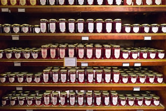 Shelves filled with jars of preserves, neatly organized in rows. The jars have gold lids and labels featuring decorative designs. The labels indicate various flavors, suggesting a variety of options.