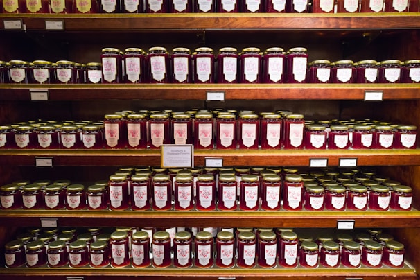Shelves filled with jars of preserves, neatly organized in rows. The jars have gold lids and labels featuring decorative designs. The labels indicate various flavors, suggesting a variety of options.