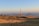 A landscape featuring a telecommunications tower on a grassy hill. Additional smaller towers and buildings are situated around the main tower. The background includes distant mountain ranges under a clear blue sky with a gradient from warm hues near the horizon.