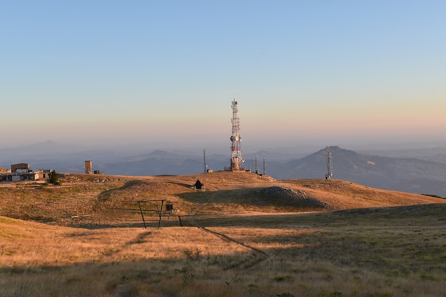 A landscape featuring a telecommunications tower on a grassy hill. Additional smaller towers and buildings are situated around the main tower. The background includes distant mountain ranges under a clear blue sky with a gradient from warm hues near the horizon.