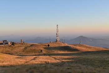 A landscape featuring a telecommunications tower on a grassy hill. Additional smaller towers and buildings are situated around the main tower. The background includes distant mountain ranges under a clear blue sky with a gradient from warm hues near the horizon.