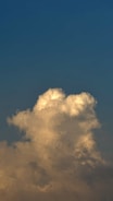 A large, fluffy cloud with hints of sunlight is set against a backdrop of a gradient blue sky, transitioning from deeper tones at the top to lighter towards the bottom.
