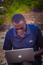 A person wearing sunglasses and a navy blue polo shirt is using a laptop outdoors. The background features blurred greenery and a stone wall, suggesting an outdoor setting.