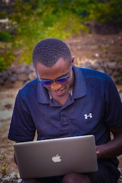 A person wearing sunglasses and a navy blue polo shirt is using a laptop outdoors. The background features blurred greenery and a stone wall, suggesting an outdoor setting.