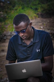 A person wearing a dark blue polo shirt and sunglasses is focused on a silver laptop with an Apple logo, set against a natural outdoor background with greenery.