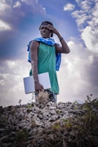 A person stands on a rocky surface, holding a laptop. They are wearing a green sleeveless shirt, a blue and white patterned scarf draped across their shoulders, and a baseball cap. The background features a dramatic, cloud-filled sky, adding a dynamic element to the scene.