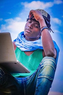 A person sitting outdoors with a laptop, dressed in casual clothing with a green tank top and blue jeans. They are wearing a hat and a light blue scarf, looking contemplatively at the laptop screen. The background is a clear sky with white clouds.