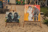 Two paintings are displayed on wooden easels outdoors. The painting on the left shows two gorillas sitting side by side, depicted in dark tones against a colorful, abstract background. The painting on the right portrays a person leaning close to another figure, both surrounded by vibrant colors including red and yellow.
