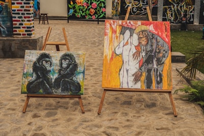 Two paintings are displayed on wooden easels outdoors. The painting on the left shows two gorillas sitting side by side, depicted in dark tones against a colorful, abstract background. The painting on the right portrays a person leaning close to another figure, both surrounded by vibrant colors including red and yellow.
