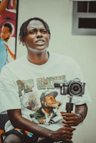 A person with braided hair wearing a T-shirt featuring a graphic print is holding a camera mounted on a stabilizer. In the background, there is a colorful mural depicting people.