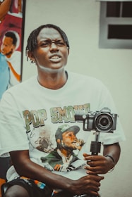 A person with braided hair wearing a T-shirt featuring a graphic print is holding a camera mounted on a stabilizer. In the background, there is a colorful mural depicting people.