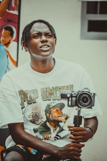 A person with braided hair wearing a T-shirt featuring a graphic print is holding a camera mounted on a stabilizer. In the background, there is a colorful mural depicting people.