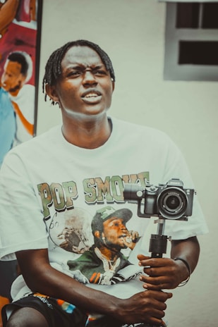 A person with braided hair wearing a T-shirt featuring a graphic print is holding a camera mounted on a stabilizer. In the background, there is a colorful mural depicting people.