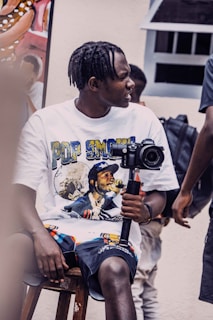 A person with braided hair sits on a wooden stool holding a camera mounted on a stabilizer. They are wearing a white t-shirt with a graphic print and dark shorts. There is another person partially visible in the background, along with a poster and some equipment.