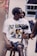 A person with braided hair sits on a wooden stool holding a camera mounted on a stabilizer. They are wearing a white t-shirt with a graphic print and dark shorts. There is another person partially visible in the background, along with a poster and some equipment.
