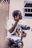A person with braided hair is sitting on a stool, holding a camera mounted on a stabilizer. The individual is wearing a graphic t-shirt featuring a musician and patterned shorts. The scene includes a painting in the background and a window to the right.