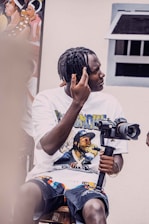 A person with braided hair is sitting on a stool, holding a camera mounted on a stabilizer. The individual is wearing a graphic t-shirt featuring a musician and patterned shorts. The scene includes a painting in the background and a window to the right.