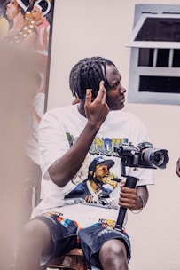 A person with braided hair is sitting on a stool, holding a camera mounted on a stabilizer. The individual is wearing a graphic t-shirt featuring a musician and patterned shorts. The scene includes a painting in the background and a window to the right.