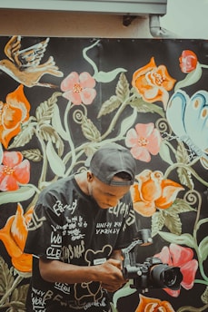A man with a black cap and a graphic t-shirt is holding a camera mounted on a stabilizer. He is positioned in front of a vibrant mural featuring large floral designs, a bird, and a butterfly, all with vivid colors like orange, pink, green, and blue.