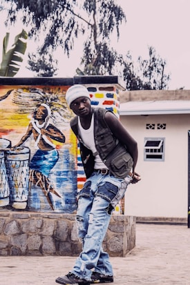 A person wearing a white beanie, a sleeveless vest, and ripped jeans stands in a courtyard, with a vibrant mural depicting musicians playing drums in the background. The mural is painted with colorful strokes and features cultural themes. The setting has a rustic exterior wall with a window.