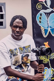 A person holding a camera mounted on a stabilizer, wearing a white t-shirt featuring graphics and text. In the background, there is a colorful mural with a large butterfly design.