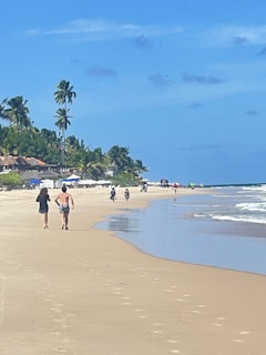 A sandy beach stretches alongside clear blue waters under a bright sky, with palm trees lining the shore. People walk along the shoreline while others appear to ride bicycles in the distance. The area is lively, with umbrellas and beachgoers in the background.