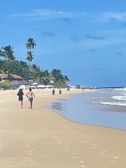 A sandy beach stretches alongside clear blue waters under a bright sky, with palm trees lining the shore. People walk along the shoreline while others appear to ride bicycles in the distance. The area is lively, with umbrellas and beachgoers in the background.