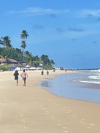 A sandy beach stretches alongside clear blue waters under a bright sky, with palm trees lining the shore. People walk along the shoreline while others appear to ride bicycles in the distance. The area is lively, with umbrellas and beachgoers in the background.