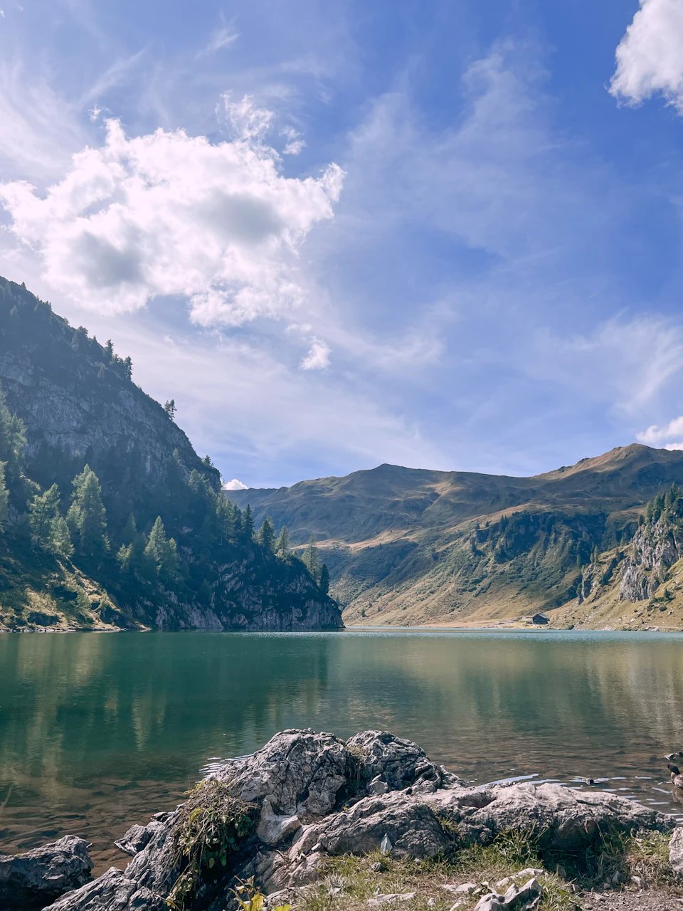 The image depicts a serene mountain lake nestled amidst lush green mountains and trees. The calm, light blue water reflects the surrounding landscape, creating a mirror-like effect that enhances the tranquility of the scene. On one side of the lake, there is a rocky shore with patches of grass and shrubs, adding a touch of ruggedness to the otherwise smooth surface of the water.

The mountains in the background are covered in trees, their green foliage contrasting beautifully against the blue sky dotted with white clouds. The image is taken from a high vantage point, providing a comprehensive view of the lake and its surroundings.