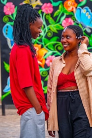 Two people are standing outdoors in front of a colorful mural with vibrant floral designs. They are looking at each other and smiling warmly, suggesting a sense of connection or friendship. The person on the left is wearing a red shirt and gray pants, while the person on the right is dressed in a red top, striped shirt, and black pants.
