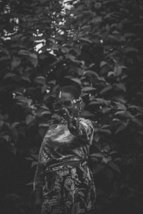 A person with braided hair and large sunglasses stands in front of a dense backdrop of foliage, wearing a patterned outfit and making a peace sign. The image is in black and white, giving it a vintage feel.
