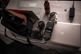 A table covered with a white cloth holds several items including a silver laptop, a digital camera with a lens attached, a lens cap, an external flash, a camera bag, a power adapter, and a beverage bottle. All objects appear casually arranged, suggesting a photography or tech-related setup.