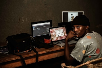 A person sits at a desk facing sideways, wearing a beanie and a shirt with polka dots. The desk holds three open laptops displaying various screens. A camera bag and other electronics are also present, creating a workspace environment with a focus on technology.