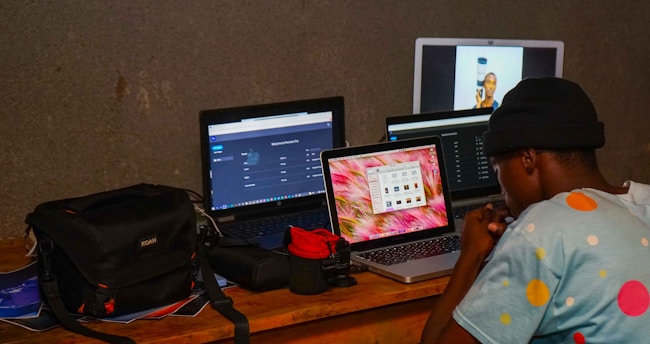 A person wearing a polka-dotted shirt and a hat is sitting at a wooden table with three laptops in front of them. The laptops display different screens, including software interfaces and an image. Several camera bags and camera equipment are placed beside the laptops.