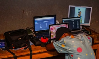 A person is leaning over a desk cluttered with multiple laptops and electronic equipment. The person is wearing a colorful polka-dotted shirt and a hat. One laptop displays a web page, while another shows an image of a person holding an object. A black bag and other tech accessories are also on the table. The background is a plain wall.