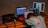 A person is leaning over a desk cluttered with multiple laptops and electronic equipment. The person is wearing a colorful polka-dotted shirt and a hat. One laptop displays a web page, while another shows an image of a person holding an object. A black bag and other tech accessories are also on the table. The background is a plain wall.