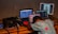 A person is leaning over a desk cluttered with multiple laptops and electronic equipment. The person is wearing a colorful polka-dotted shirt and a hat. One laptop displays a web page, while another shows an image of a person holding an object. A black bag and other tech accessories are also on the table. The background is a plain wall.