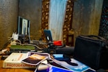 A cluttered desk displaying photography equipment, including a computer monitor with an image on the screen, a laptop with an illuminated logo, a camera with a lens, assorted cables, photos scattered across the surface, and a book titled 'Photographer's'. The background features a textured wall and partially visible curtains with floral patterns.