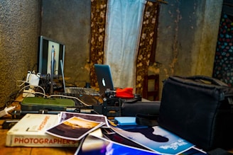 A cluttered desk displaying photography equipment, including a computer monitor with an image on the screen, a laptop with an illuminated logo, a camera with a lens, assorted cables, photos scattered across the surface, and a book titled 'Photographer's'. The background features a textured wall and partially visible curtains with floral patterns.