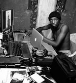 A person is seated at a cluttered workspace with multiple laptops and photography equipment. The setting appears to be inside a room with patterned curtains and a rough-textured wall. Various camera lenses, photos, and gadgets are scattered across the wooden table. The individual is looking directly at the camera, wearing a beanie and sitting shirtless.