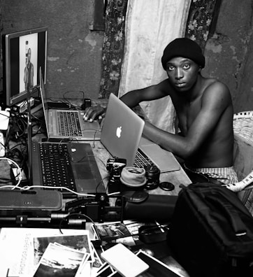 A person is seated at a cluttered workspace with multiple laptops and photography equipment. The setting appears to be inside a room with patterned curtains and a rough-textured wall. Various camera lenses, photos, and gadgets are scattered across the wooden table. The individual is looking directly at the camera, wearing a beanie and sitting shirtless.