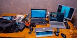 A wooden desk cluttered with electronic devices and photography equipment including two laptops, a third laptop partially visible beneath them, a digital camera, various camera lenses, bags, and cables. A separate monitor displays an image of a person holding a camera lens. Several photography magazines are also scattered on the desk.