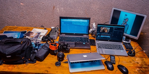A wooden desk cluttered with electronic devices and photography equipment including two laptops, a third laptop partially visible beneath them, a digital camera, various camera lenses, bags, and cables. A separate monitor displays an image of a person holding a camera lens. Several photography magazines are also scattered on the desk.
