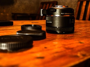 A camera lens and a few lens caps rest on a polished wooden table. The lens is a compact model with a shiny finish, situated in a warm ambient light setting. The background is slightly out of focus, highlighting wooden chairs and adding depth.
