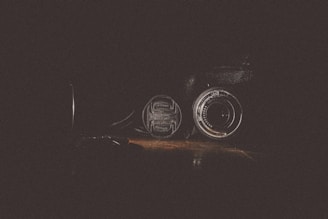A camera lens placed on a wooden surface with dramatic lighting that highlights its metallic and glass components against a dark background.