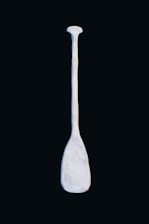 A solitary, light-colored paddle against a dark background. The paddle appears to be made of a material resembling stone or ceramic, with a slightly rough texture and some markings or indentations on its surface.
