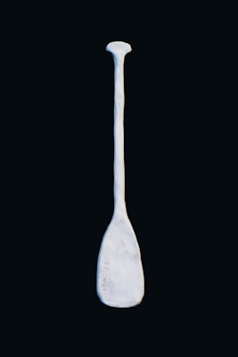 A solitary, light-colored paddle against a dark background. The paddle appears to be made of a material resembling stone or ceramic, with a slightly rough texture and some markings or indentations on its surface.